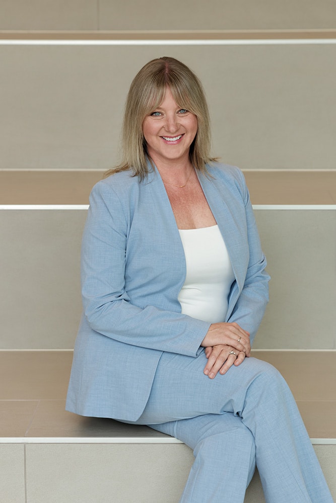 Woman in a light blue suit sitting on beige steps, smiling at the camera.