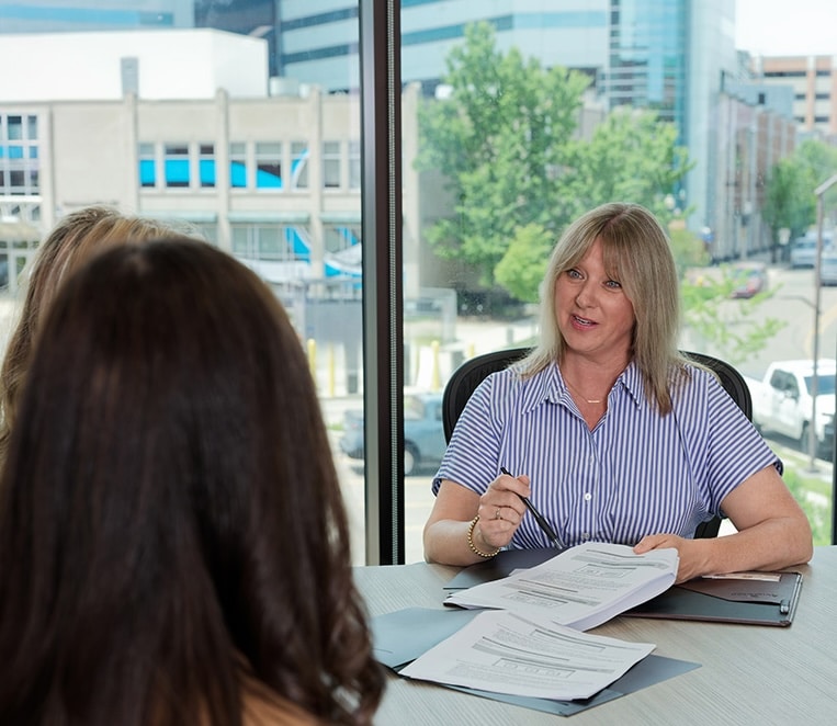 A woman holding a pen speaks with two others at a table with paperwork in an office with large windows.