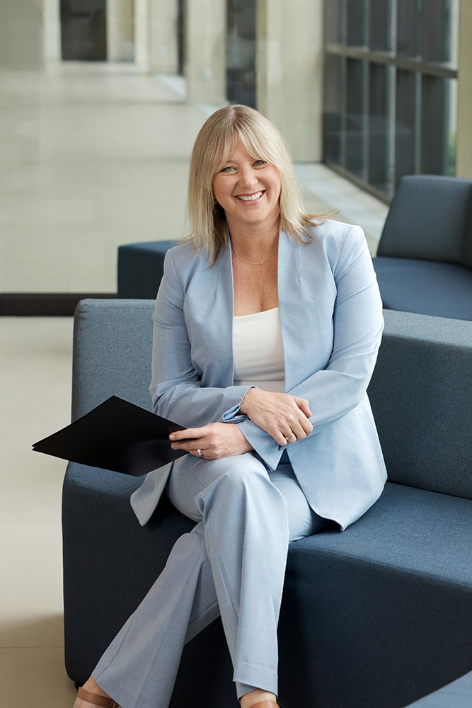 Woman in a light blue suit sits on a couch, smiling and holding a black folder in an office setting.